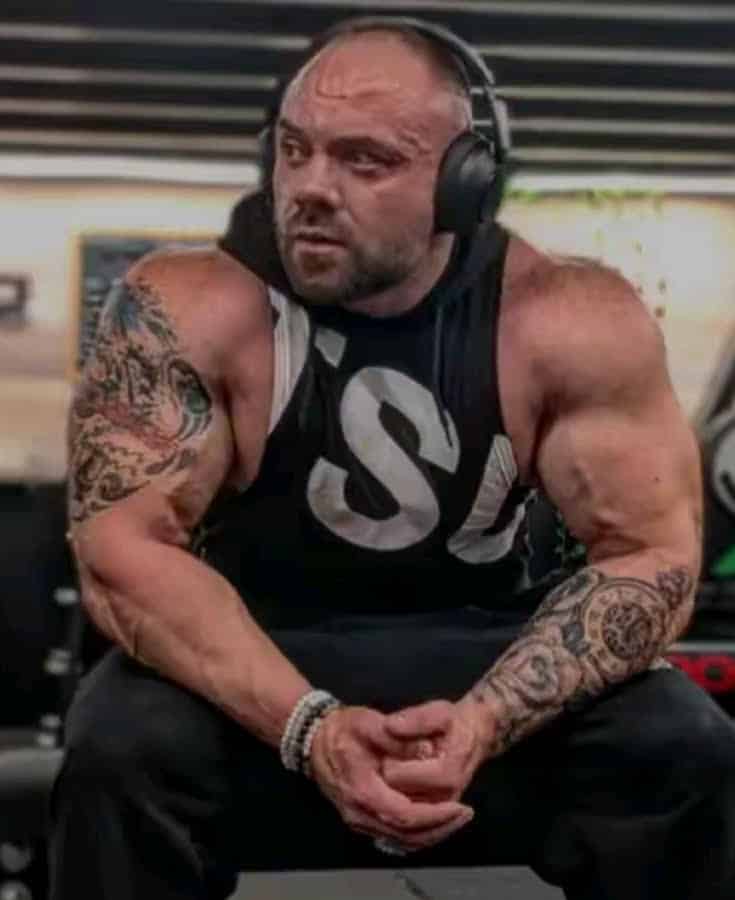 Bodybuilder musclé tatoué écoutant de la musique avec un casque, vêtu d'un débardeur noir avec un grand 'S' blanc.