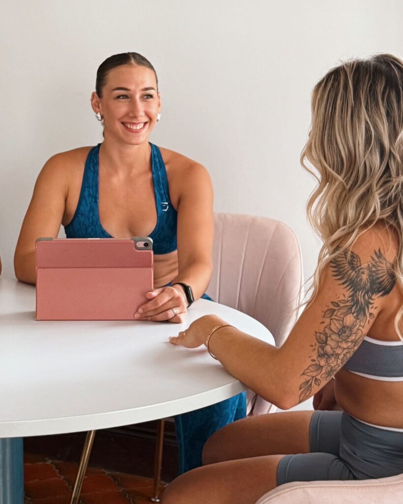 Deux femmes en tenue de sport discutent autour d'une table blanche ; l'une tient une tablette rose, l'autre a un tatouage floral.
