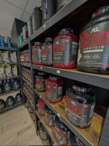 Étalage d'un magasin de suppléments : pots de protéine AEL ISO Protein (rouge et noir), shakers SKILL and NUTRITION, et sachets de protéines Isolate.