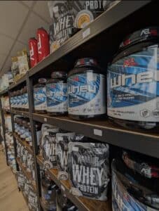 Étagères remplies de suppléments de protéines en poudre Whey ASL et Warrior, avec des bouteilles rouges et des étiquettes de prix visibles.