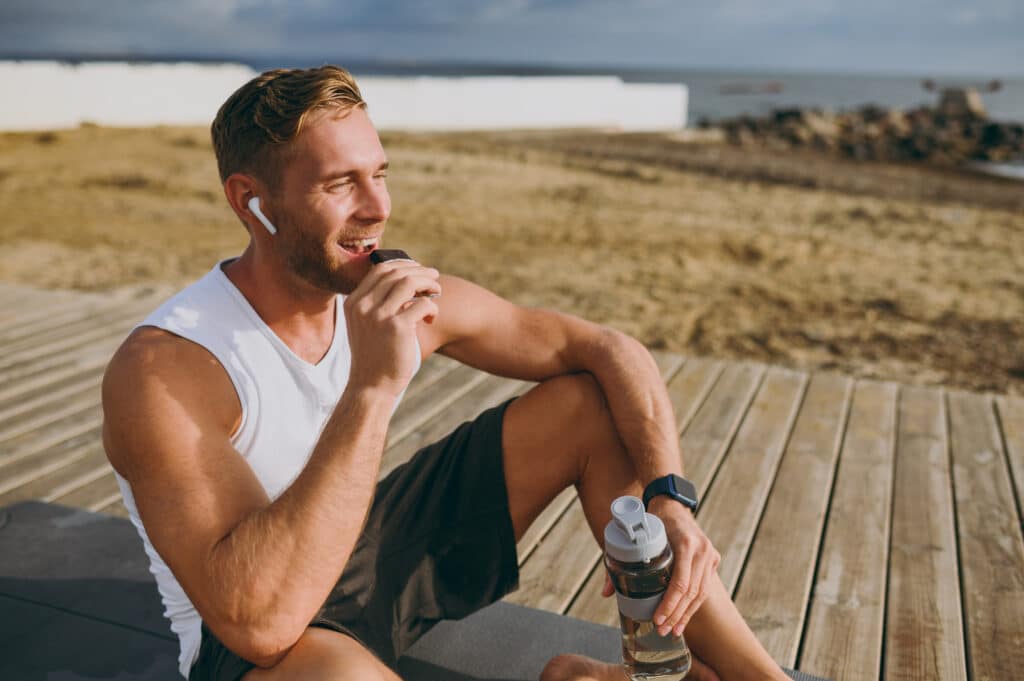 Homme athlétique souriant mange une barre protéinée sur une terrasse en bois près de la mer, tenant une gourde.