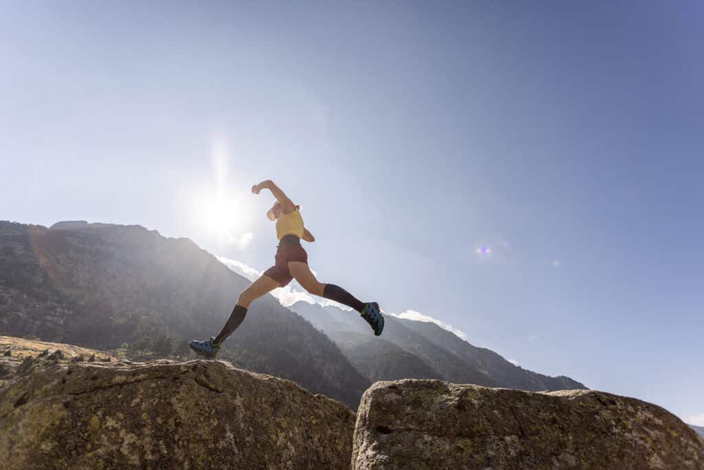 Trail runner en plein saut entre deux rochers, silhouette dynamique face au soleil éclatant et aux montagnes.